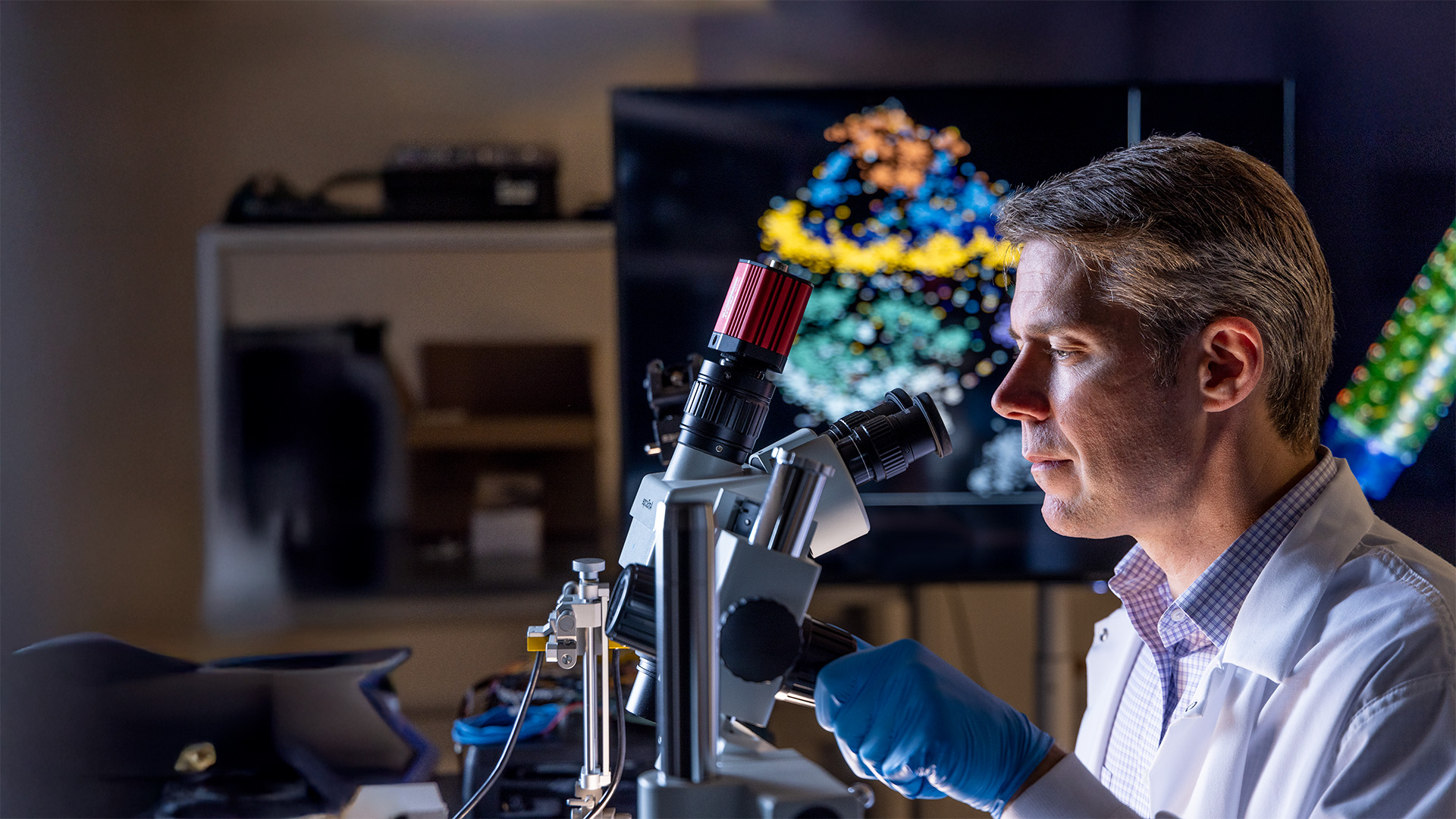 UTHealth Houston researcher in a lab coat and gloves uses a microscope while colorful molecular imaging appears on monitors in the background, representing biomedical research, scientific discovery, and advanced laboratory technology.