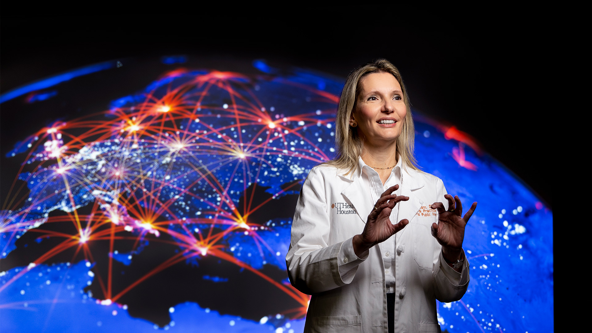 Healthcare professional in a UTHealth Houston lab coat gestures while speaking in front of a large illuminated digital globe with global network connections, representing innovation, research, technology, and international collaboration.