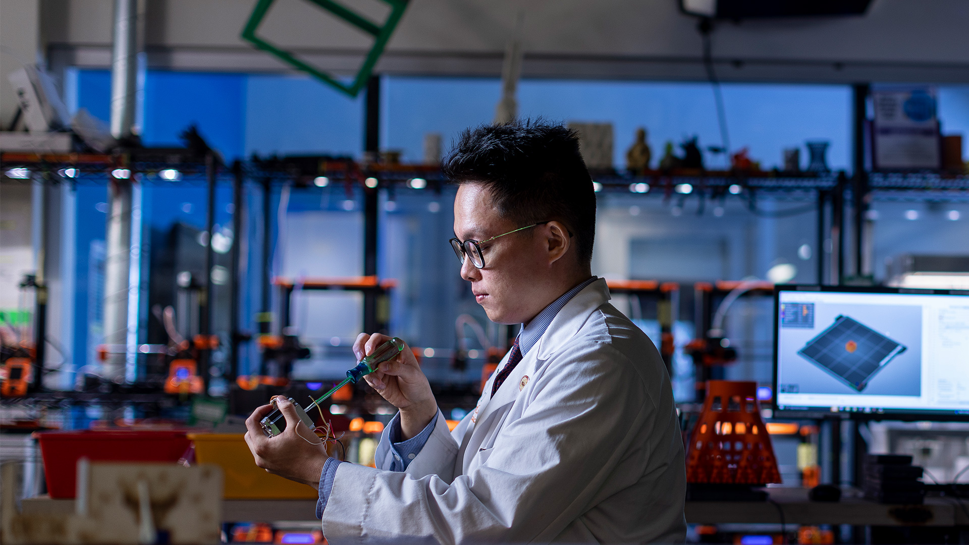 UTHealth Houston researcher in a lab coat uses a precision tool to assemble or test an electronic device in an advanced engineering laboratory, with 3D printers and computer modeling software visible in the background.