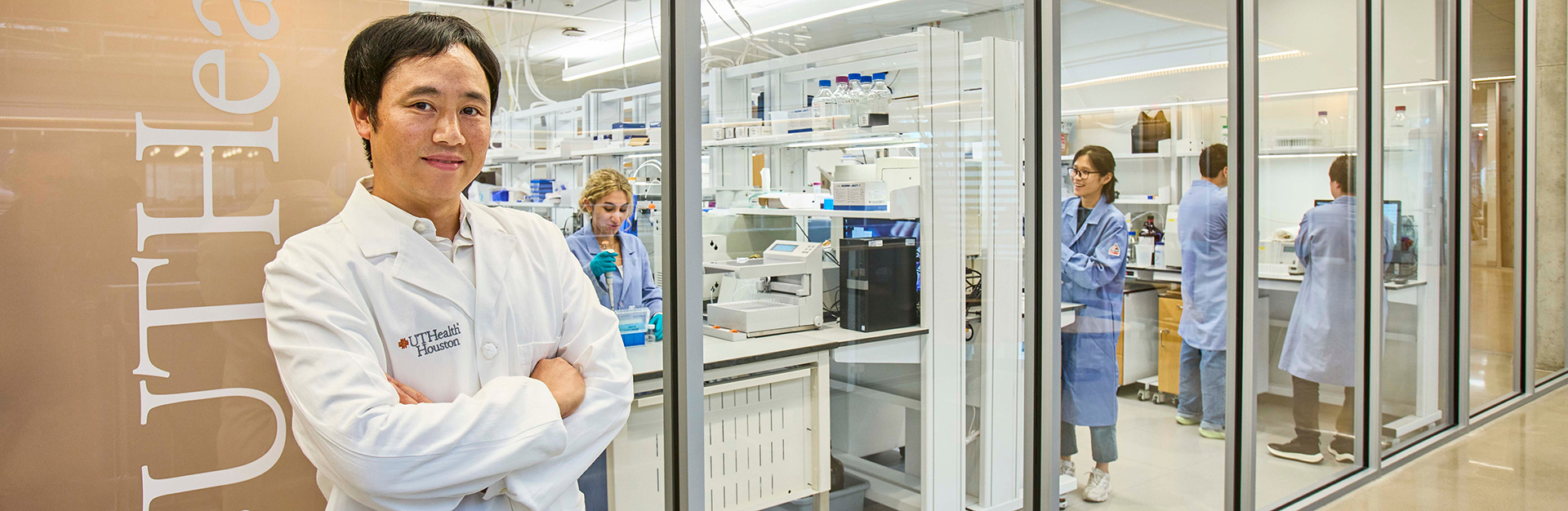 A researcher in a white UTHealth Houston lab coat stands with arms crossed in the foreground, smiling at the camera, while several colleagues work in a bright, modern laboratory behind glass walls, using scientific equipment and conducting experiments.