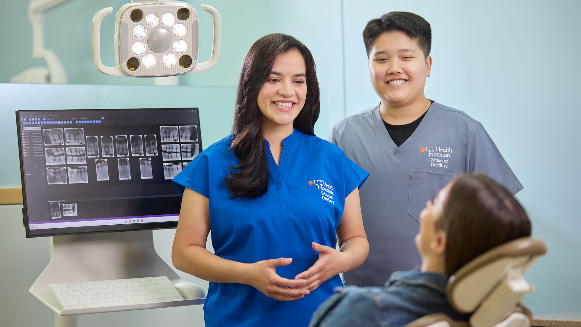 Dental students speaking with a patient beside a computer monitor in a clinical dental setting.