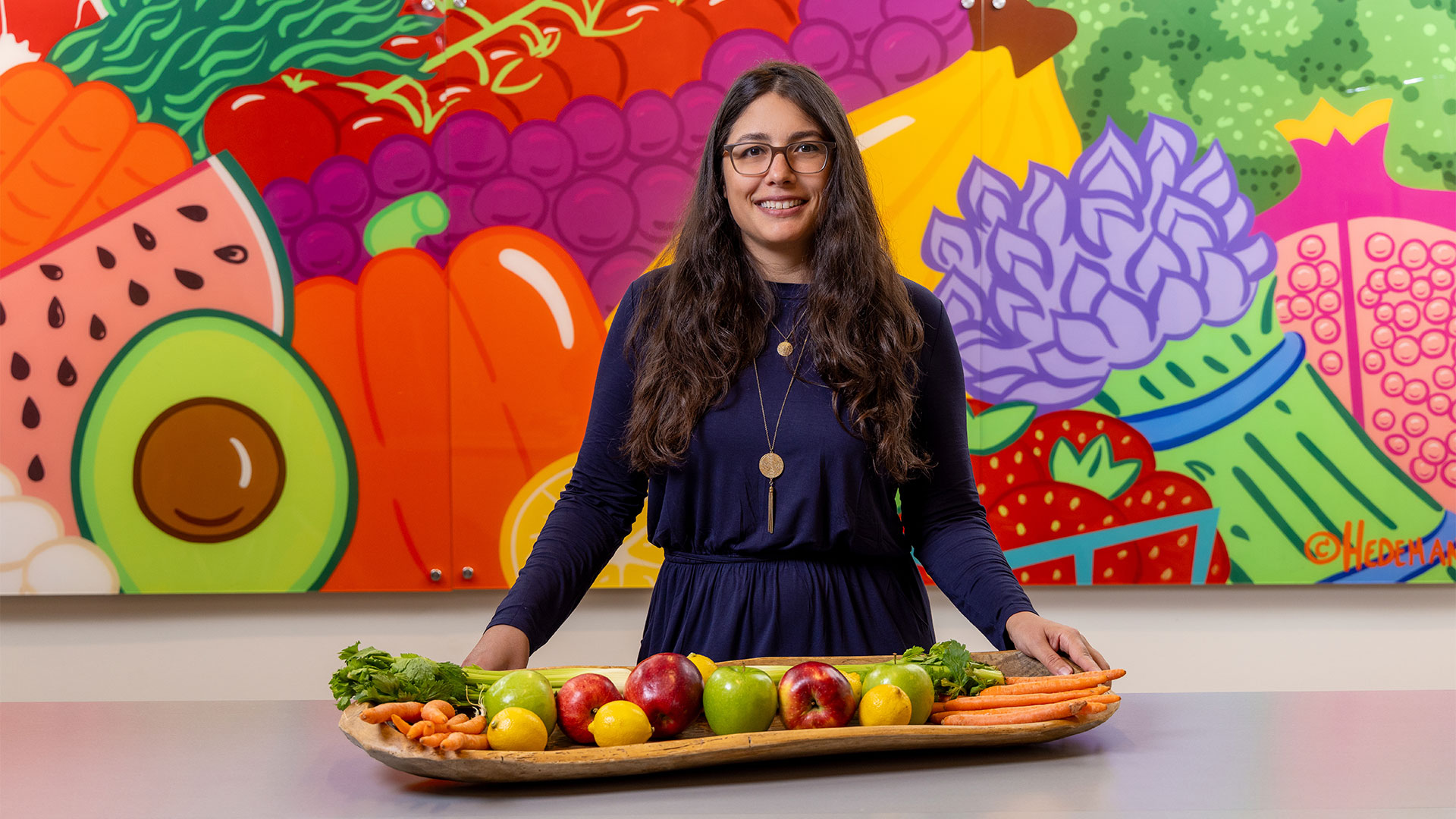 Public health student standing behind fresh fruits and vegetables with a colorful nutrition-themed mural in the background.