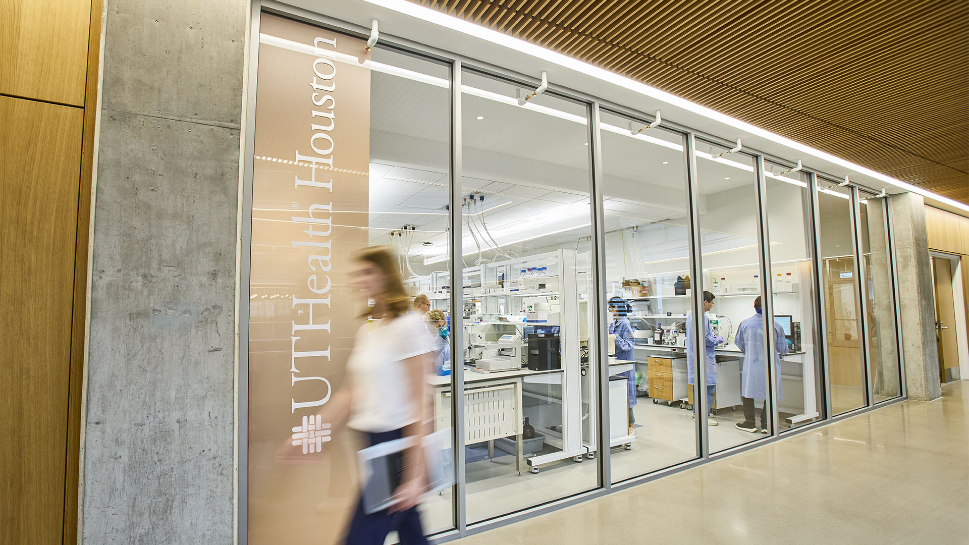 Glass-walled UTHealth Houston research laboratory with scientists working at benches and lab equipment. A blurred person walks through the hallway outside, highlighting an active biomedical research environment.