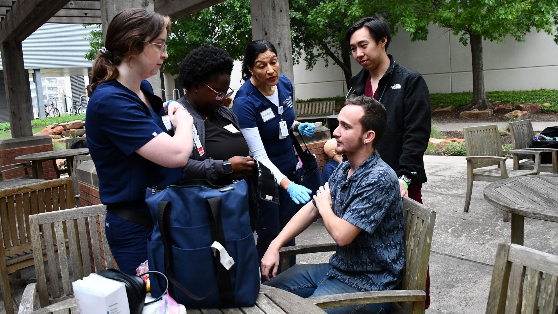 Health care trainees in scrubs practicing a patient assessment with a seated man in an outdoor courtyard