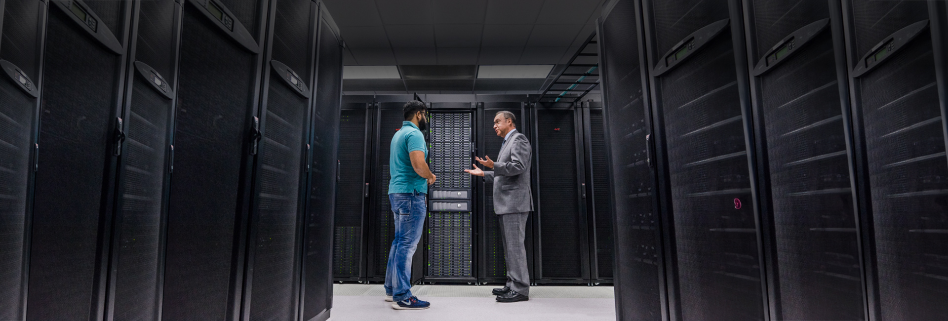 Two men talking in a server room. The man on the left is wearing a teal polo and blue jeans. The man on the right is wearing a gray suit.
