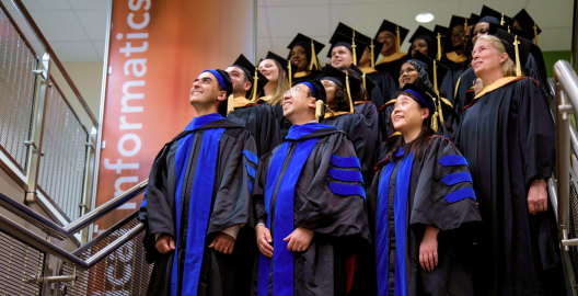 Students dressed in graduation gowns and shown in a 3/4 view smile upwards. In the background, there is an orange banner than says 'Informatics'. The studenats are standing on a staircase.