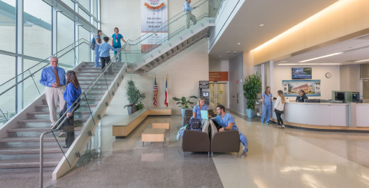 Students and faculty gathered in UTHealth Houston School of Dentistry. There is a large staircase on the left with people standing and chatting. In the middle, there are students sitting on gray benches.