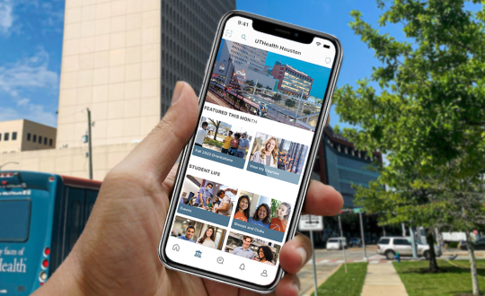 A hand holds a phone with the UTHealth Student mobile app open. In the background, there is the UTHealth Houston University Center Tower, a UTHealth Houston Bus, a bright blue sky, and a tree.