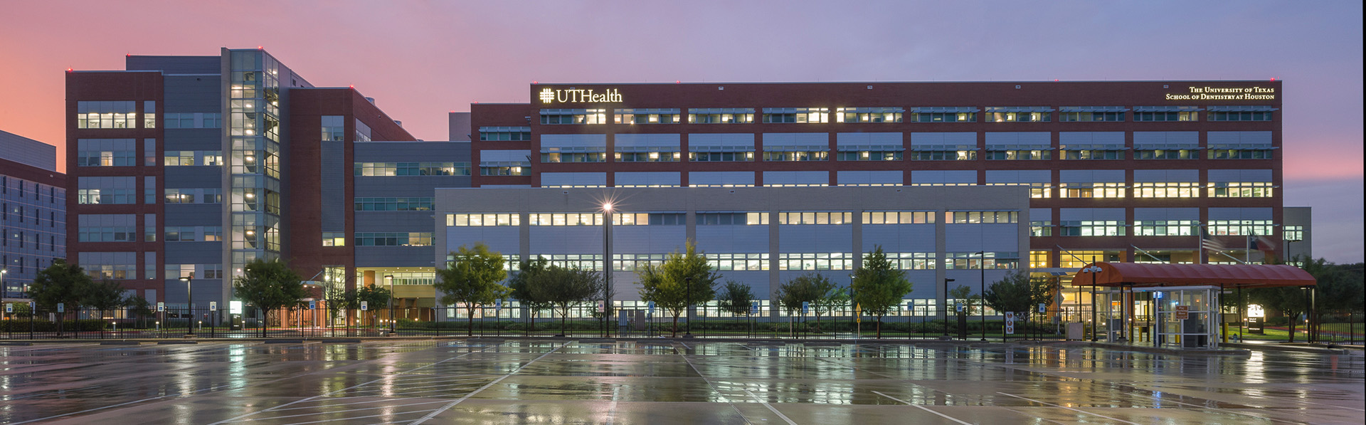 Evening view of the UTHealth Houston School of Dentistry building with illuminated signage, red and gray exterior, and an empty parking lot in the foreground.