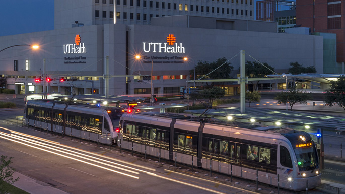 Evening view of UTHealth Houston’s University Center Tower building with light rail train passing in front and illuminated signage on the building’s facade.
