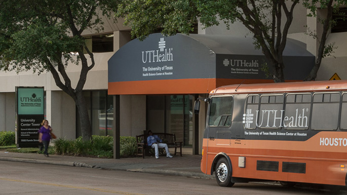 Awning entrance at UTHealth Houston’s University Center Tower with a branded UTHealth Houston shuttle bus parked in front and people waiting nearby.