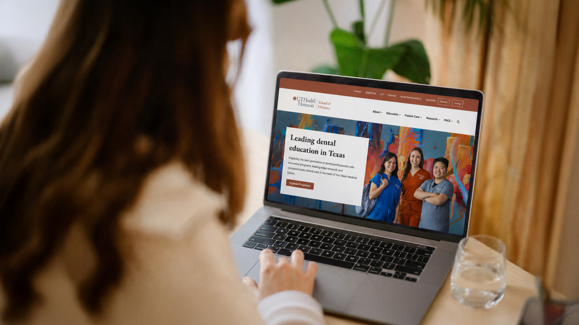 A person uses a laptop displaying the UTHealth Houston School of Dentistry website while sitting at a table. A glass of water and houseplants are nearby.