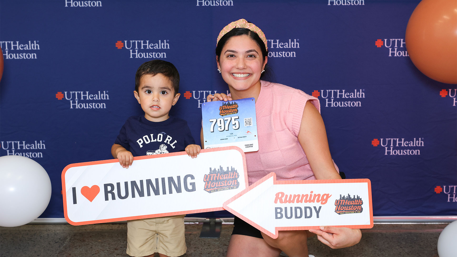 A smiling woman and a young child pose in front of a UTHealth Houston backdrop at a running event. The woman holds a race bib and a sign reading 'Running Buddy,' while the child holds a sign that says 'I love Running.' Balloons are visible on either side.