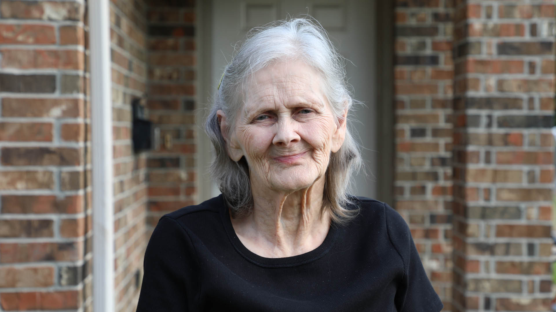 An older woman with gray hair stands in front of a brick house, facing the camera with a slight smile. She is wearing a black shirt. A doorway and brick columns are visible in the background