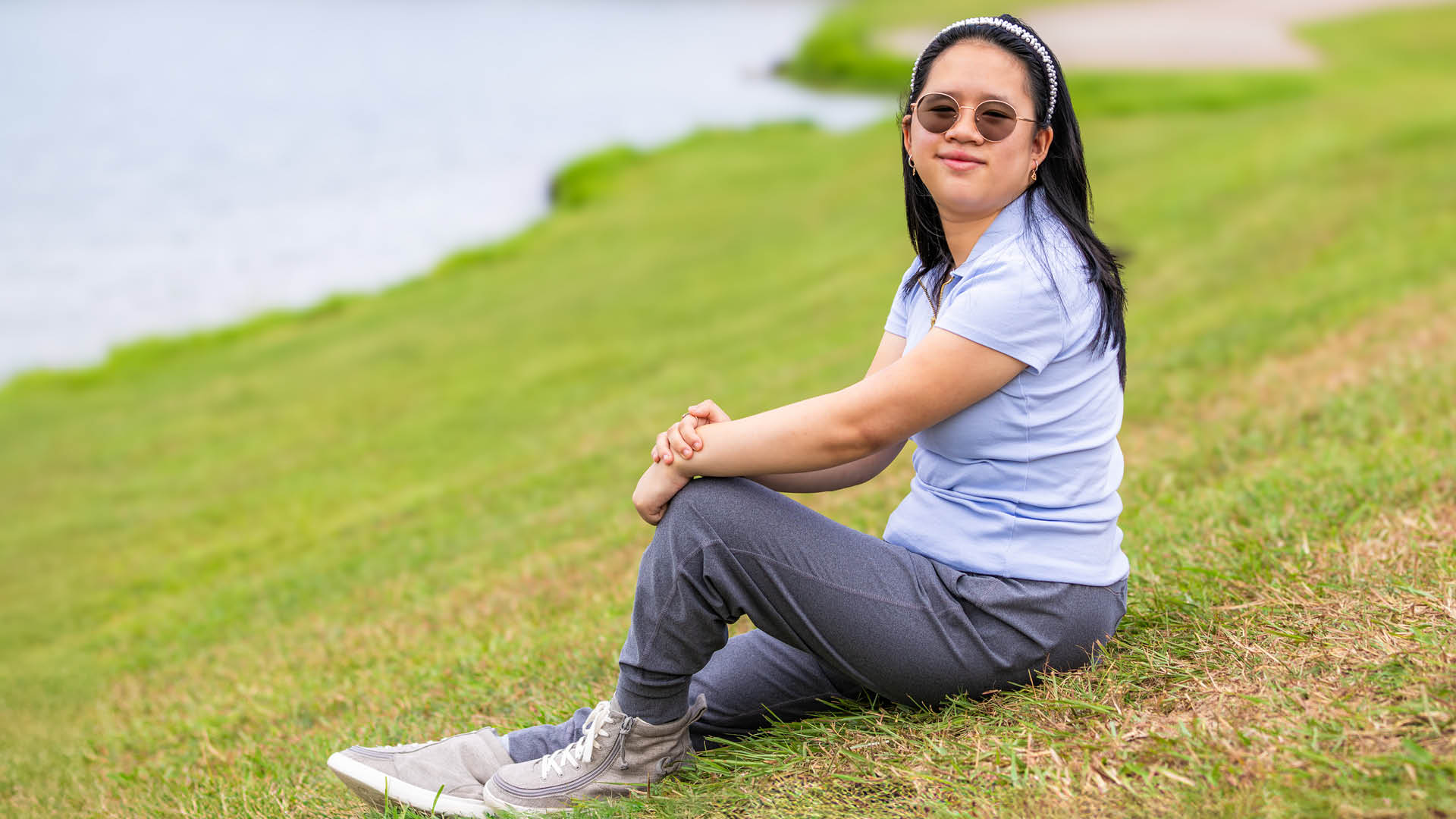 A person sits on a grassy slope near the water, facing the camera. They are wearing sunglasses, a light blue shirt, gray pants, and gray sneakers