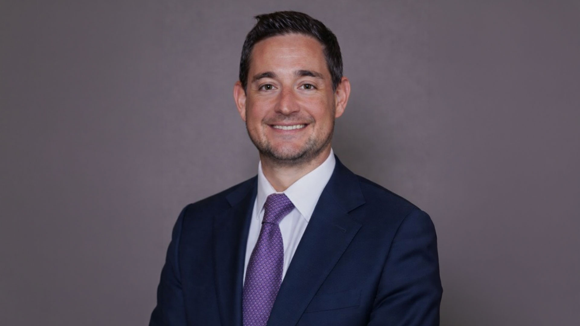 A man in a blue suit with a purple tie and white collar shirt.