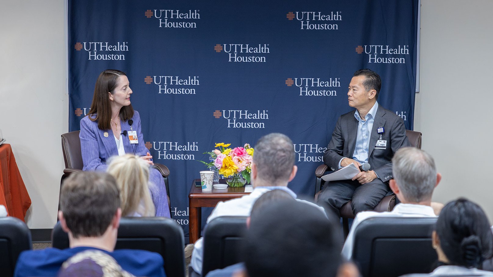 Two speakers seated in chairs during a UTHealth Houston panel discussion, with a branded backdrop and audience visible.