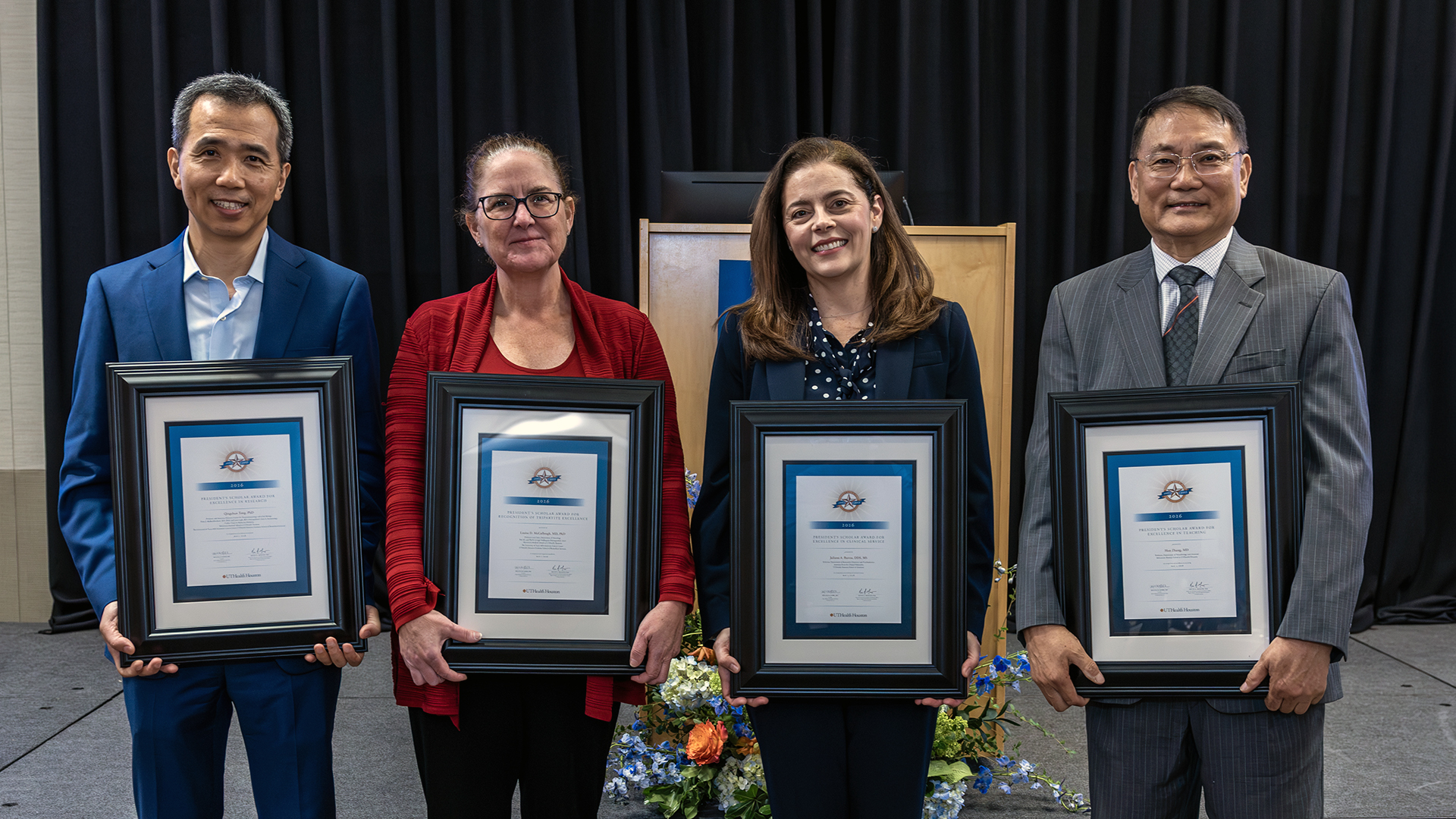 Four individuals stand in front of a stage backdrop, each holding a framed award certificate during a recognition event.