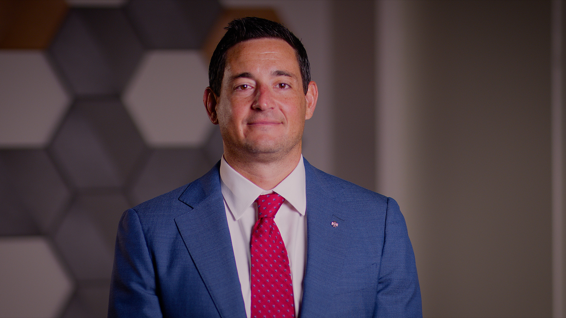Professional head-and-shoulders portrait of a person wearing a blue suit jacket, white dress shirt, and bright red patterned tie.