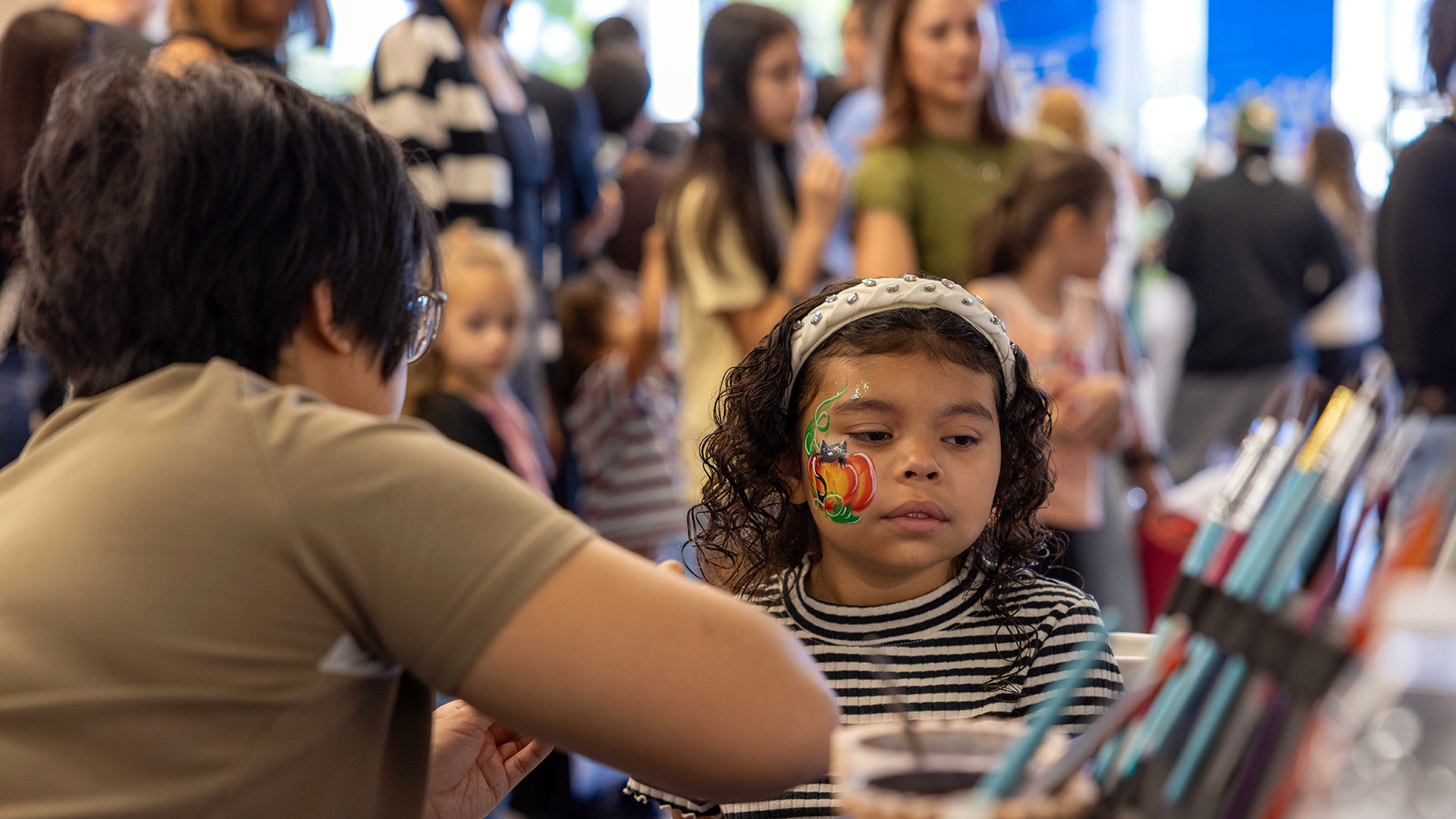 Child with a colorful pumpkin painted on her cheek sits at a face-painting station during the health fair.