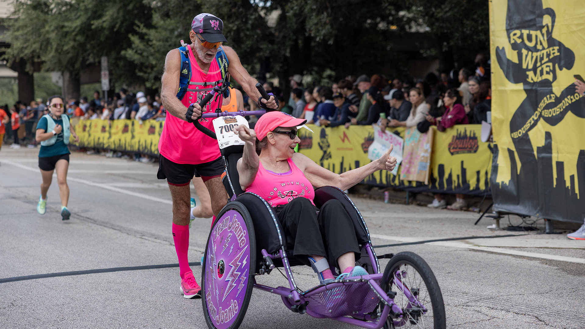 Runner pushes a racing wheelchair as the seated participant gives a thumbs-up along the Houston Half Marathon course.