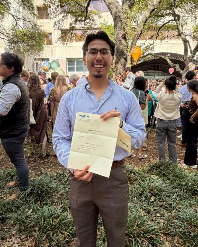 Juan Garcia smiling and holding his Match Day envelope outdoors with classmates gathered behind him.