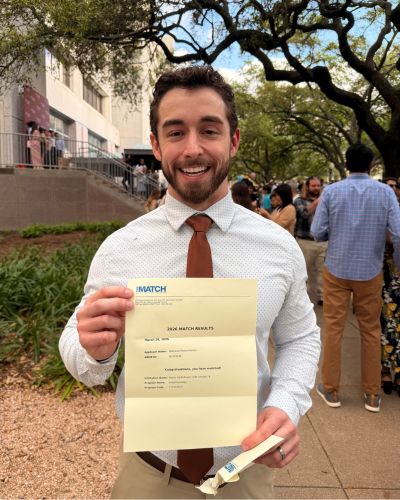 Nathaniel Hunter smiling and holding his Match Day envelope while standing outdoors with other students in the background.