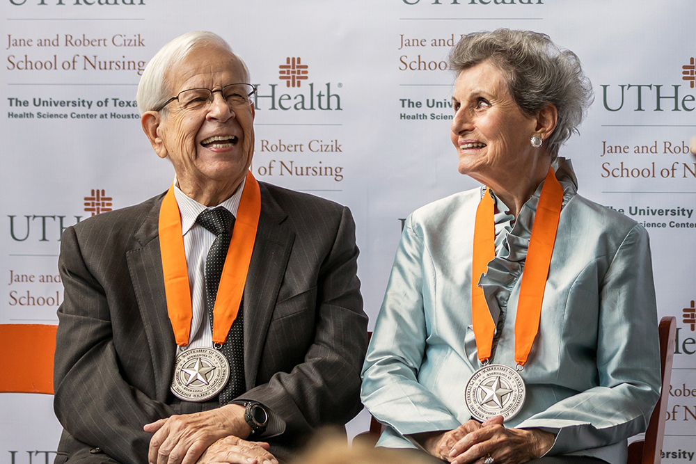 Jane and Robert Cizik wearing orange-ribbon medallions sit and smile on stage in front of a UTHealth Houston School of Nursing backdrop during a recognition ceremony.