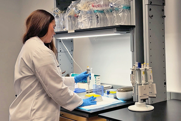 Researcher in a white lab coat and blue gloves pipetting at a laboratory bench under task lighting, with tubes and equipment organized around the workspace.