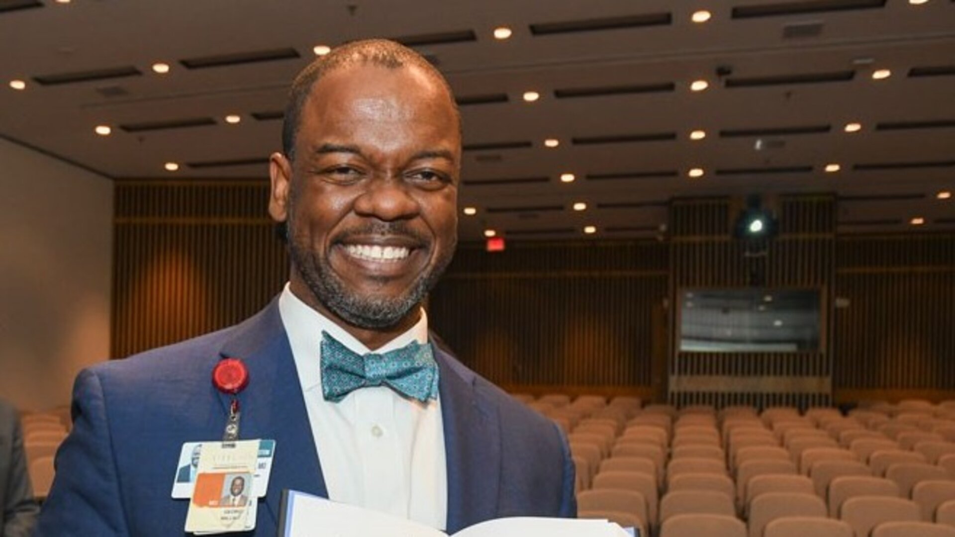 Smiling man in a blue suit and patterned bow tie holding an open book in a lecture hall with rows of empty seats, wearing an ID badge.
