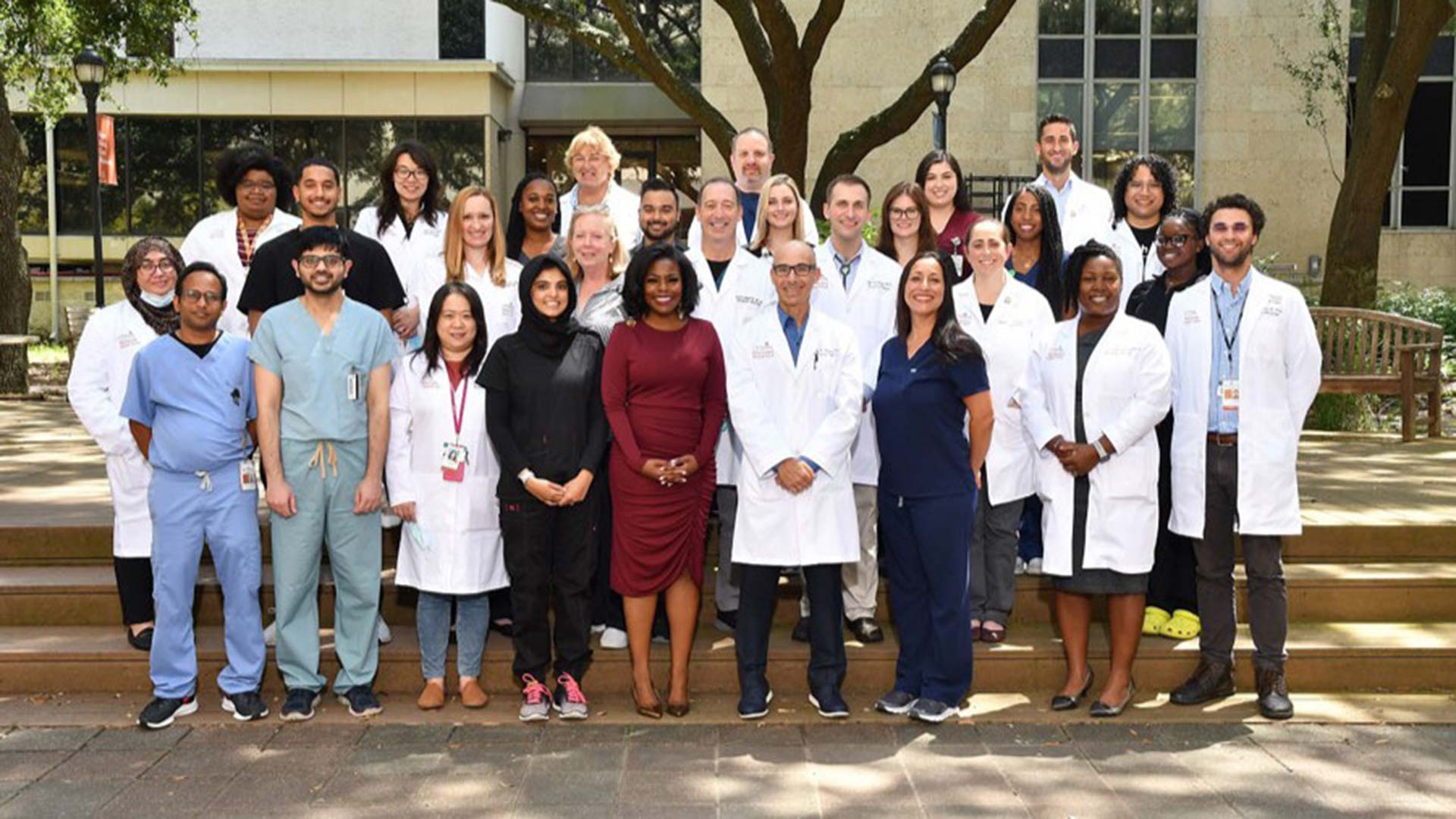 Large group of clinicians and staff in lab coats, scrubs, and business attire pose for a group photo on outdoor steps beneath trees.