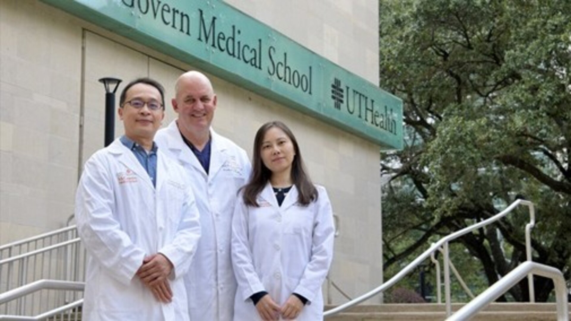 Three researchers in white lab coats standing on the steps outside McGovern Medical School at UTHealth Houston, posing near the building sign with trees in the background.