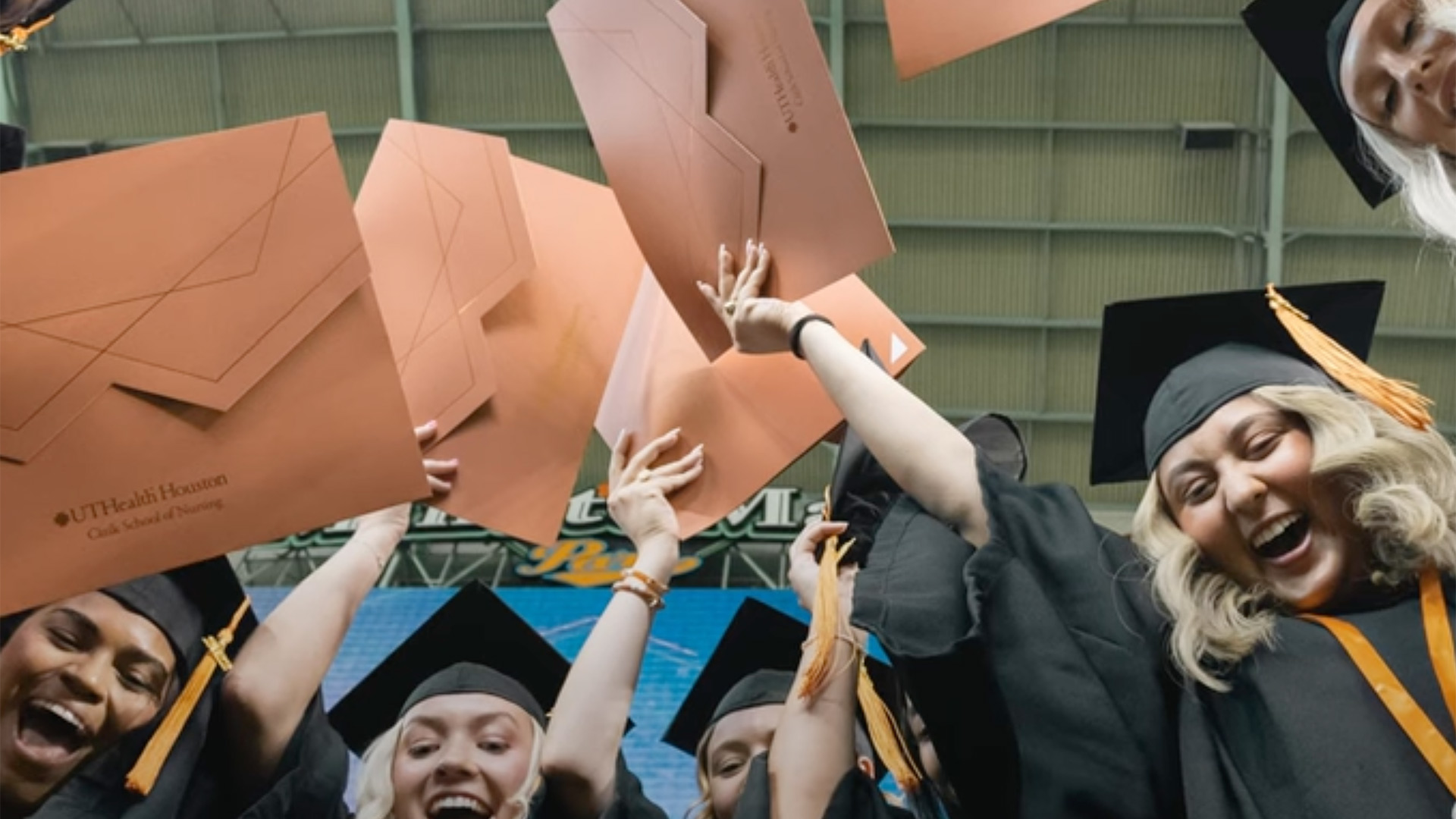 Graduates from UTHealth Houston Cizik School of Nursing celebrate in caps and gowns, raising their diplomas and smiling during commencement inside an indoor venue.