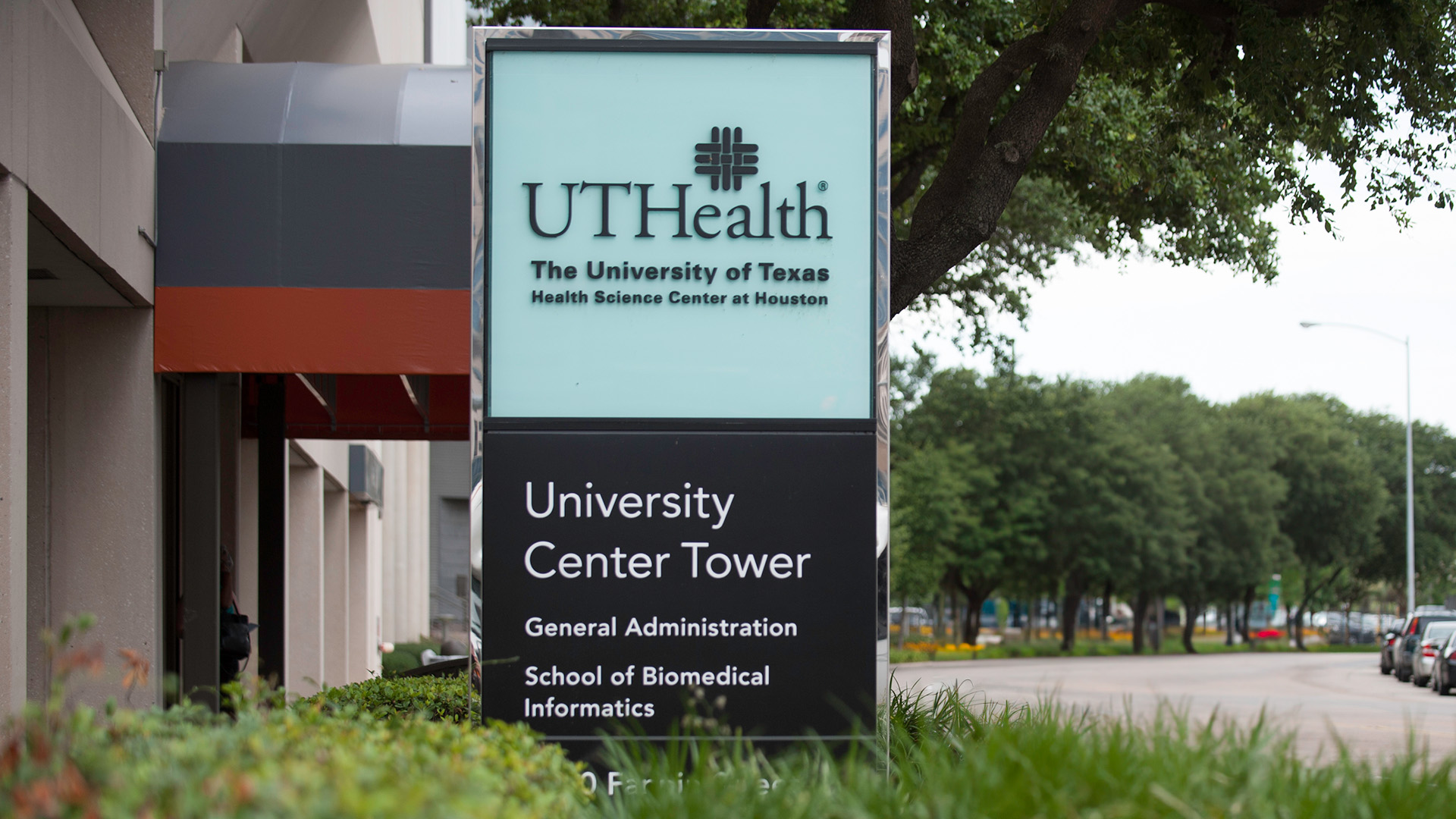 Exterior signage for UTHealth Houston's University Center Tower, showing directional information for General Administration and School of Biomedical Informatics, with cars and trees lining the adjacent street.